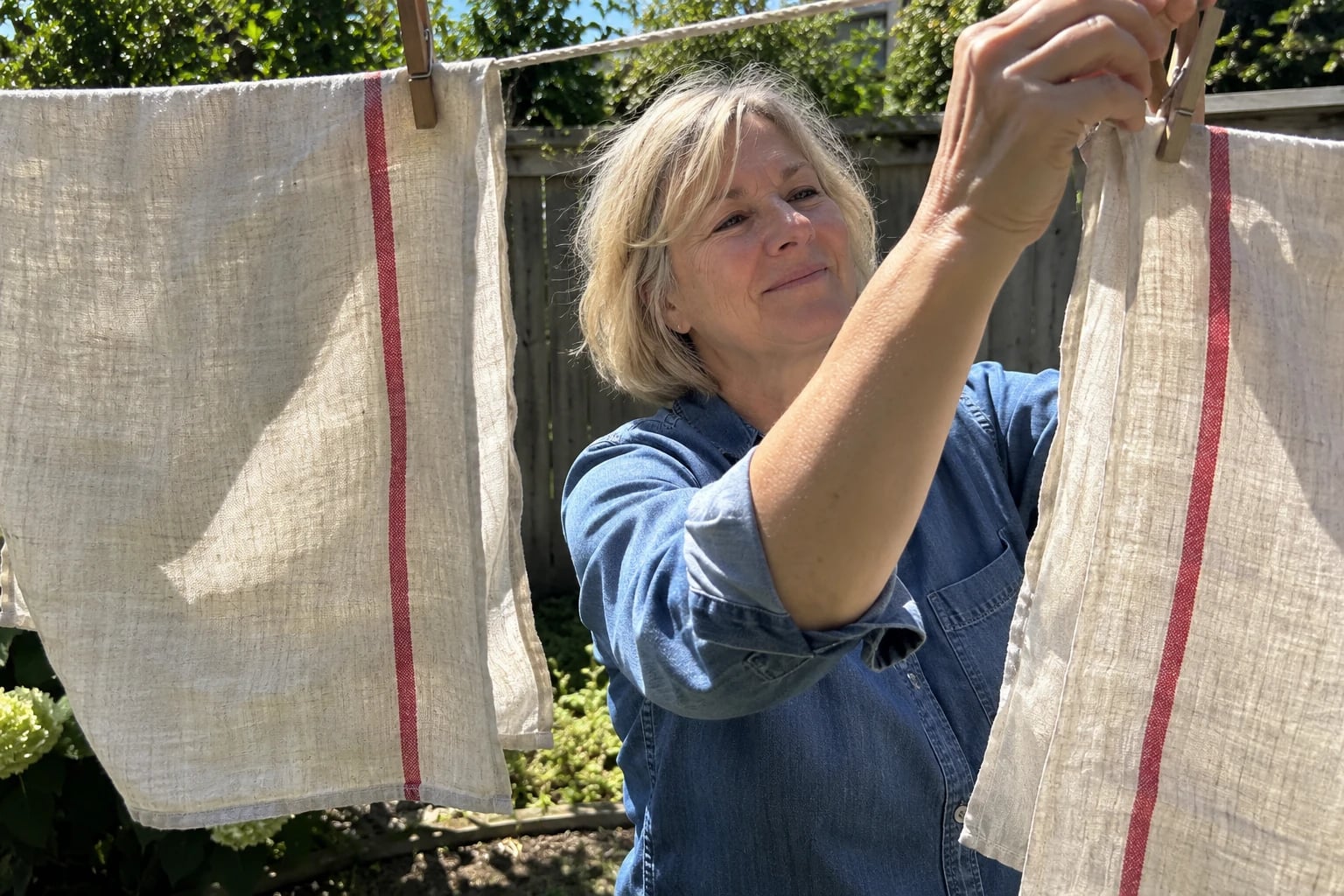 Two flat-woven natural linen tea towels with a single red stripe hang from wooden pegs on a backyard clothesline in breezy daylight, as a woman in her fifties pins up the second one.