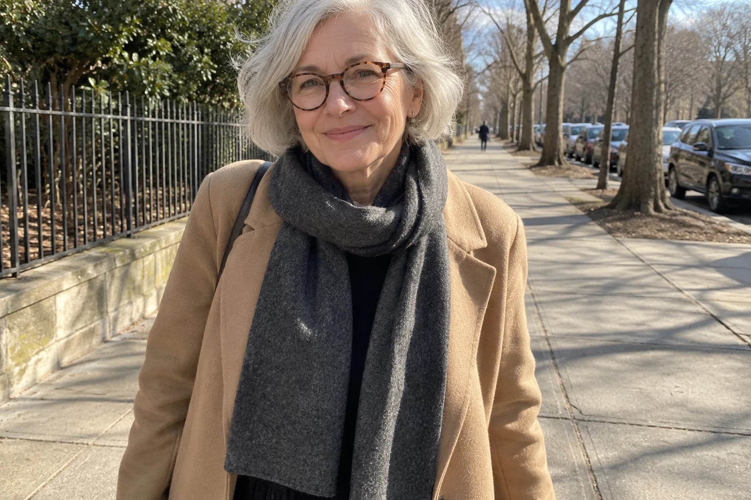 A gray-haired woman in tortoiseshell glasses wears a charcoal lambswool scarf looped over a camel coat outdoors.