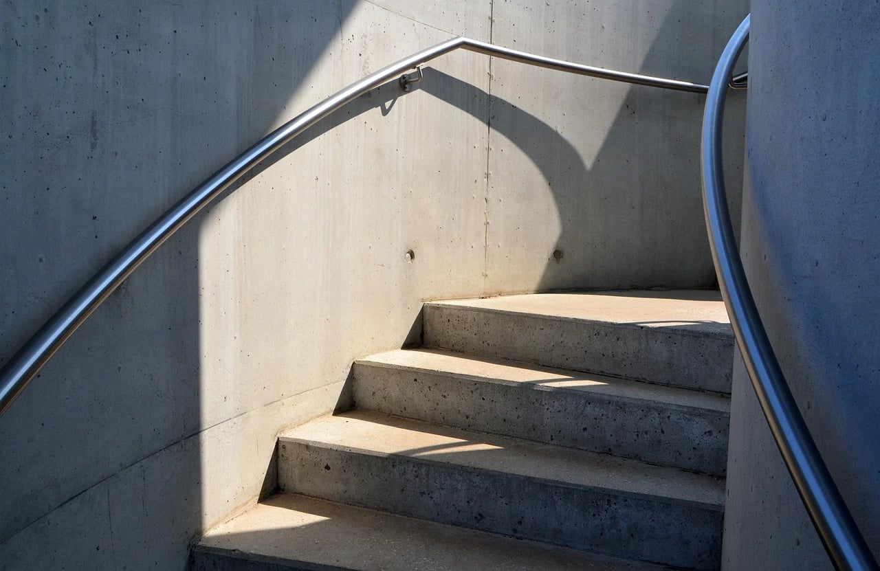 Curved concrete staircase in sunlight with sturdy stainless steel handrails along the wall and outer edge