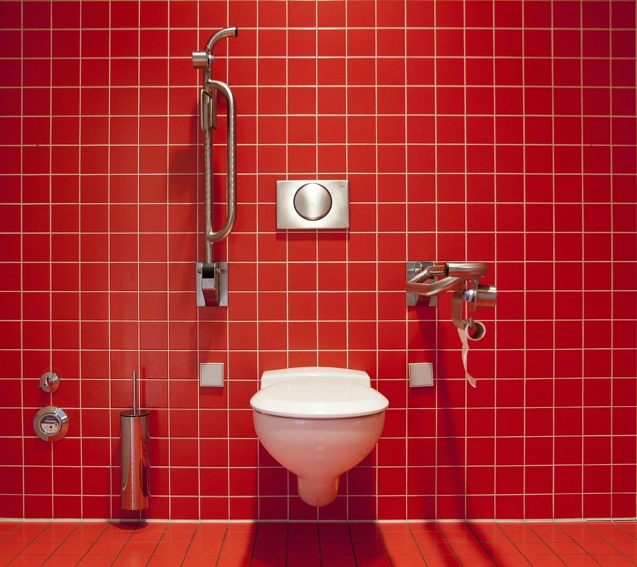 Wall-mounted white toilet with flip-up support bars on both sides against bright red tiled bathroom wall