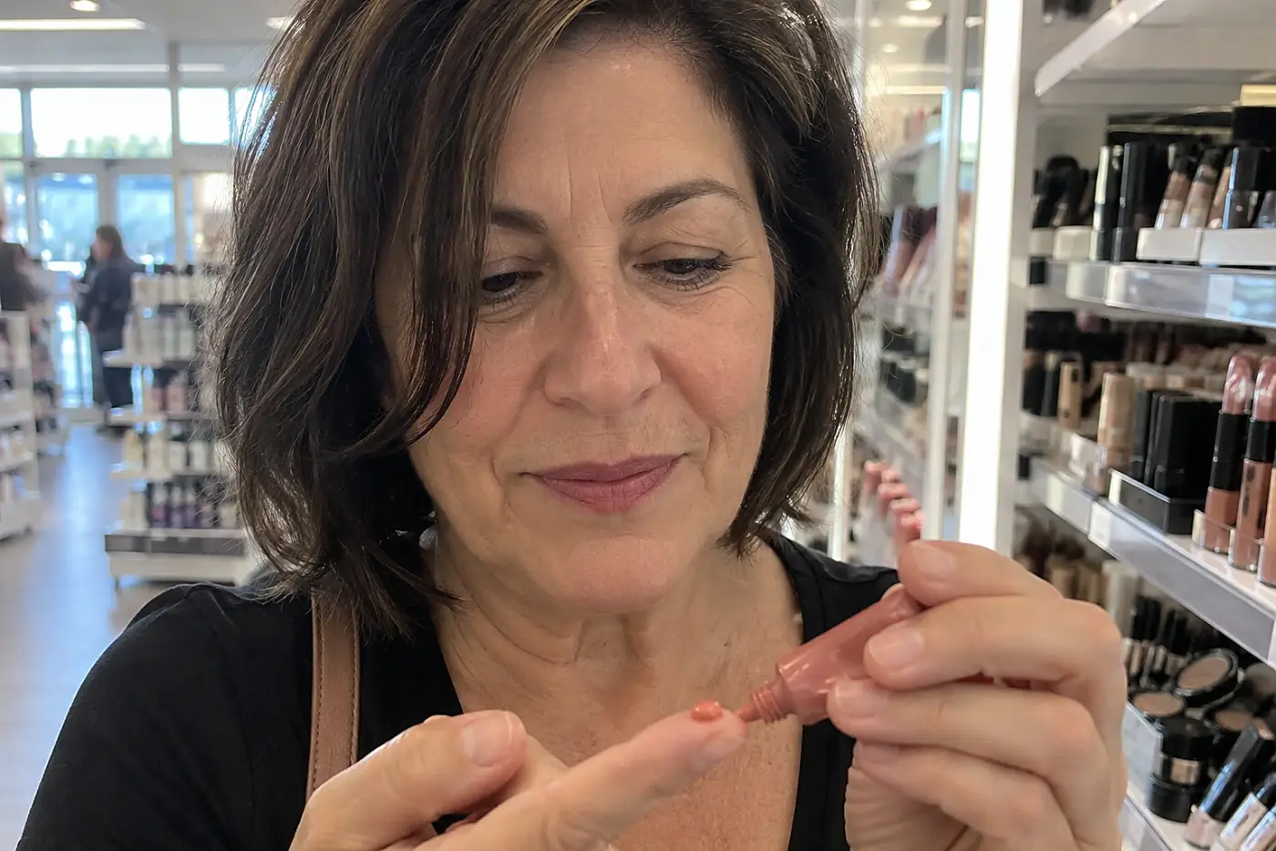Woman squeezing gel-cream blush onto a fingertip beside the mirror in a drugstore makeup aisle.
