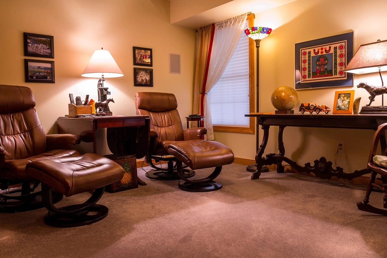 Two brown leather recliner armchairs with matching ottomans in a warmly lit living room with lamps and a desk