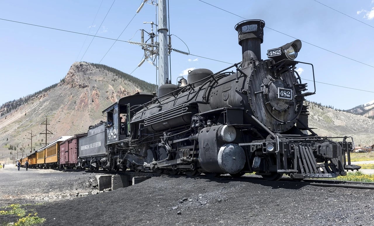 Black Durango & Silverton steam locomotive number 482 pulling vintage yellow coaches below a mountain on a sunny day