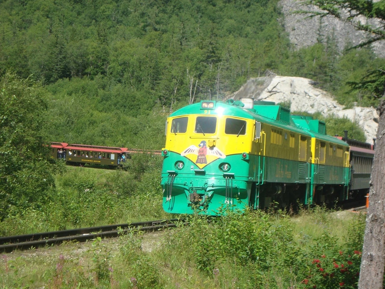 Green and yellow White Pass and Yukon Route locomotive pulling vintage passenger cars through forested mountains with a curving track