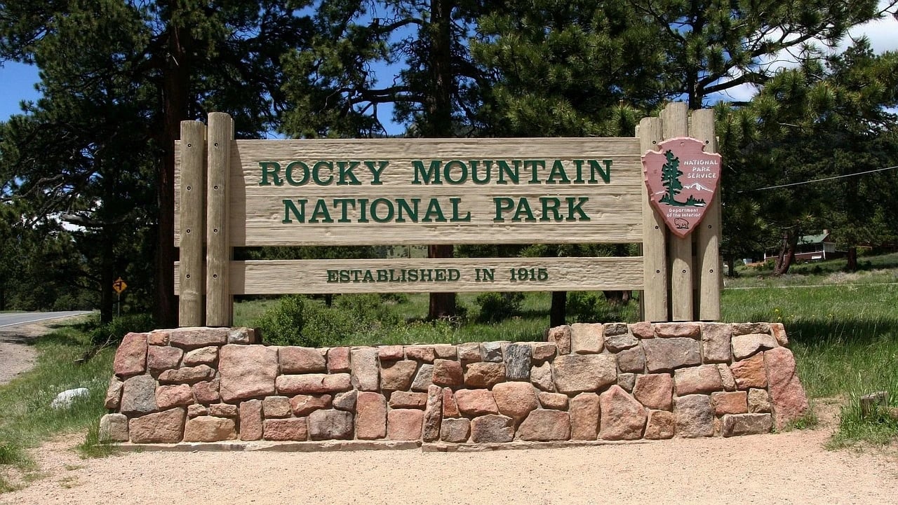 Wooden Rocky Mountain National Park entrance sign on a stone base with National Park Service arrowhead, pine trees behind
