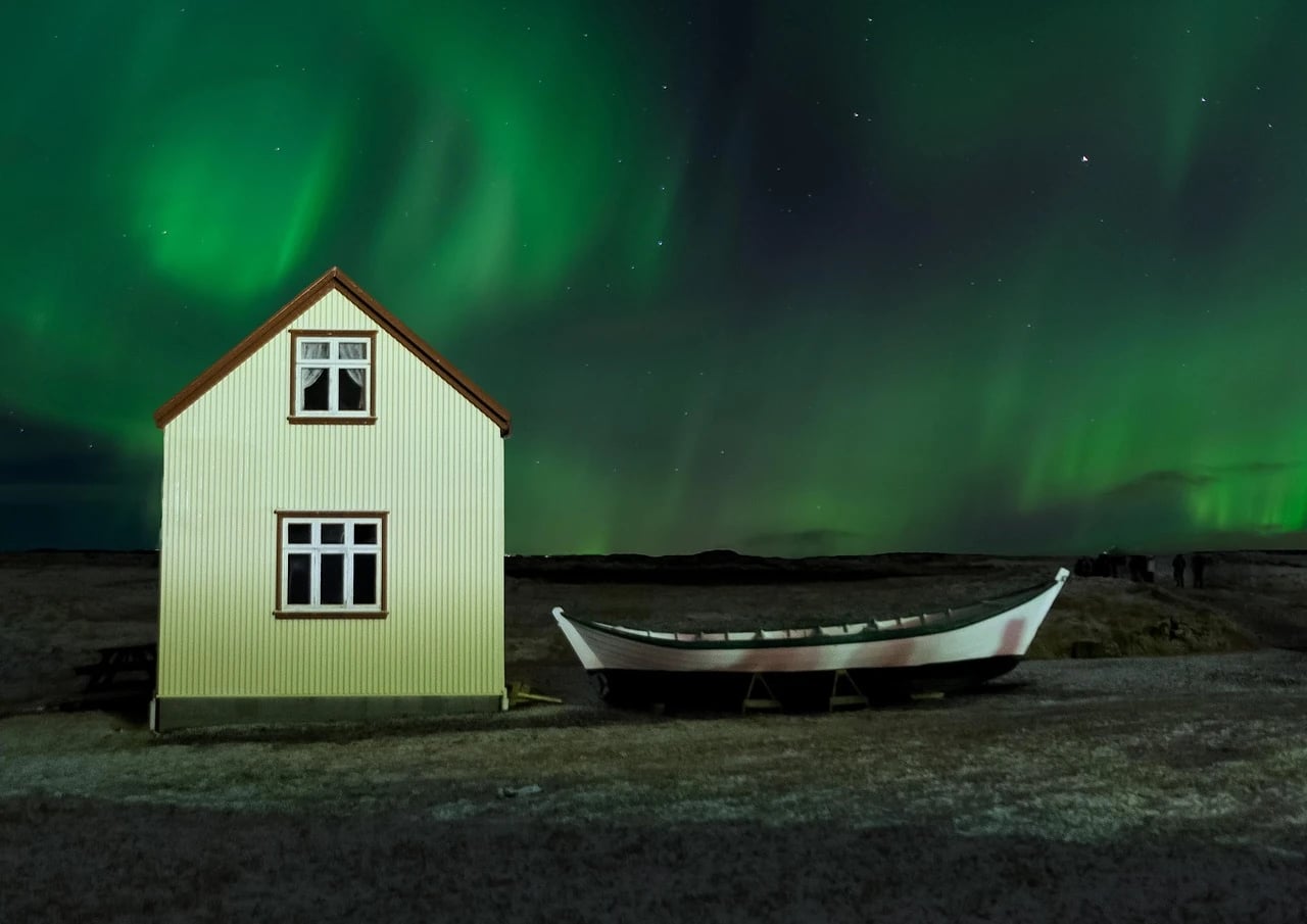Green Northern Lights glowing over a cream corrugated house and a white wooden boat on Icelandic ground