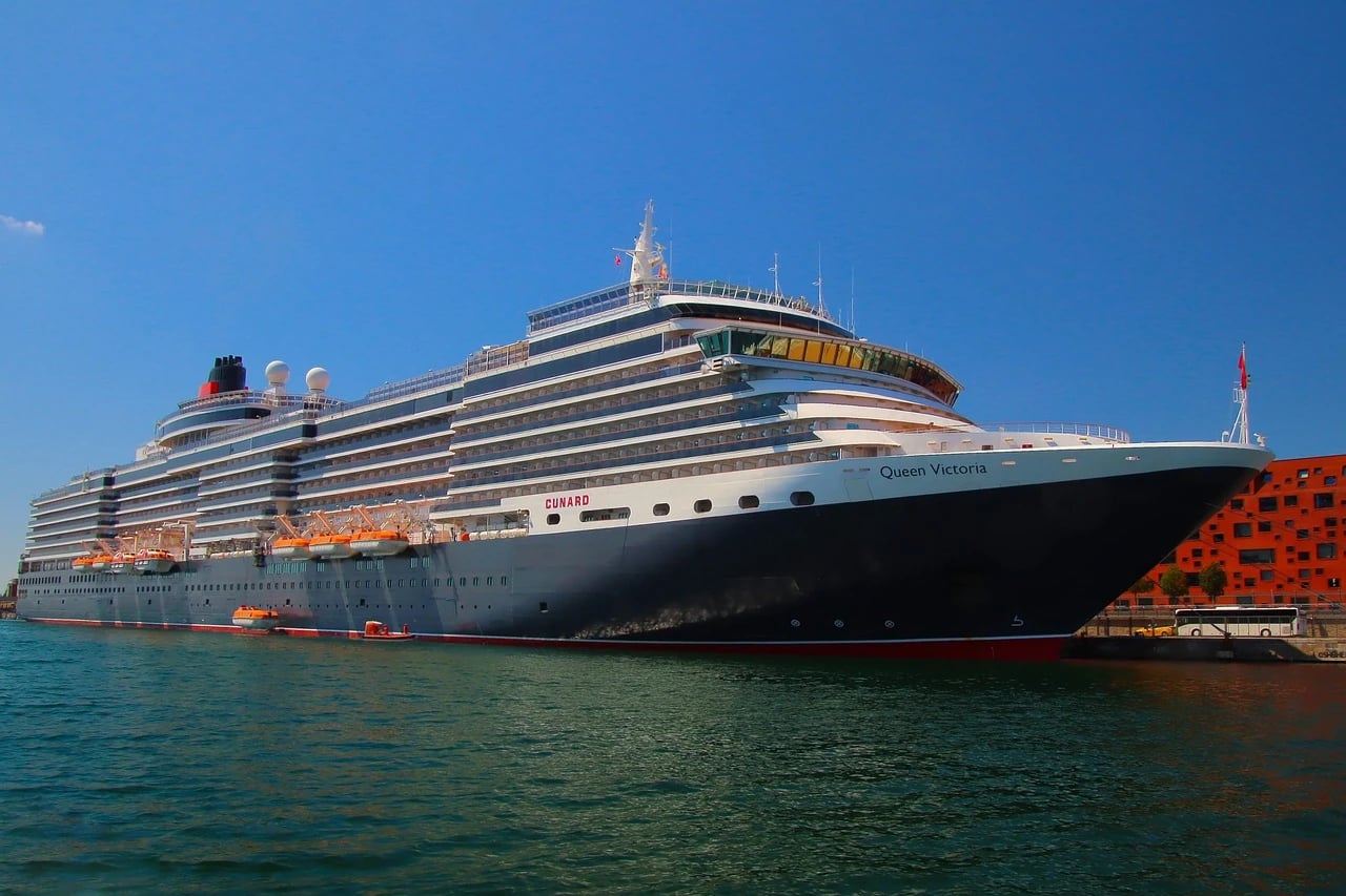 Cunard liner Queen Victoria docked in port under a clear blue sky beside an orange building