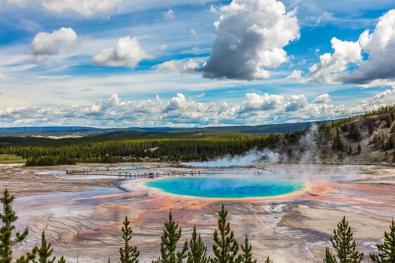 Grand Prismatic Spring in Yellowstone with turquoise water, orange edges, steam, boardwalk visitors and a cloudy blue sky