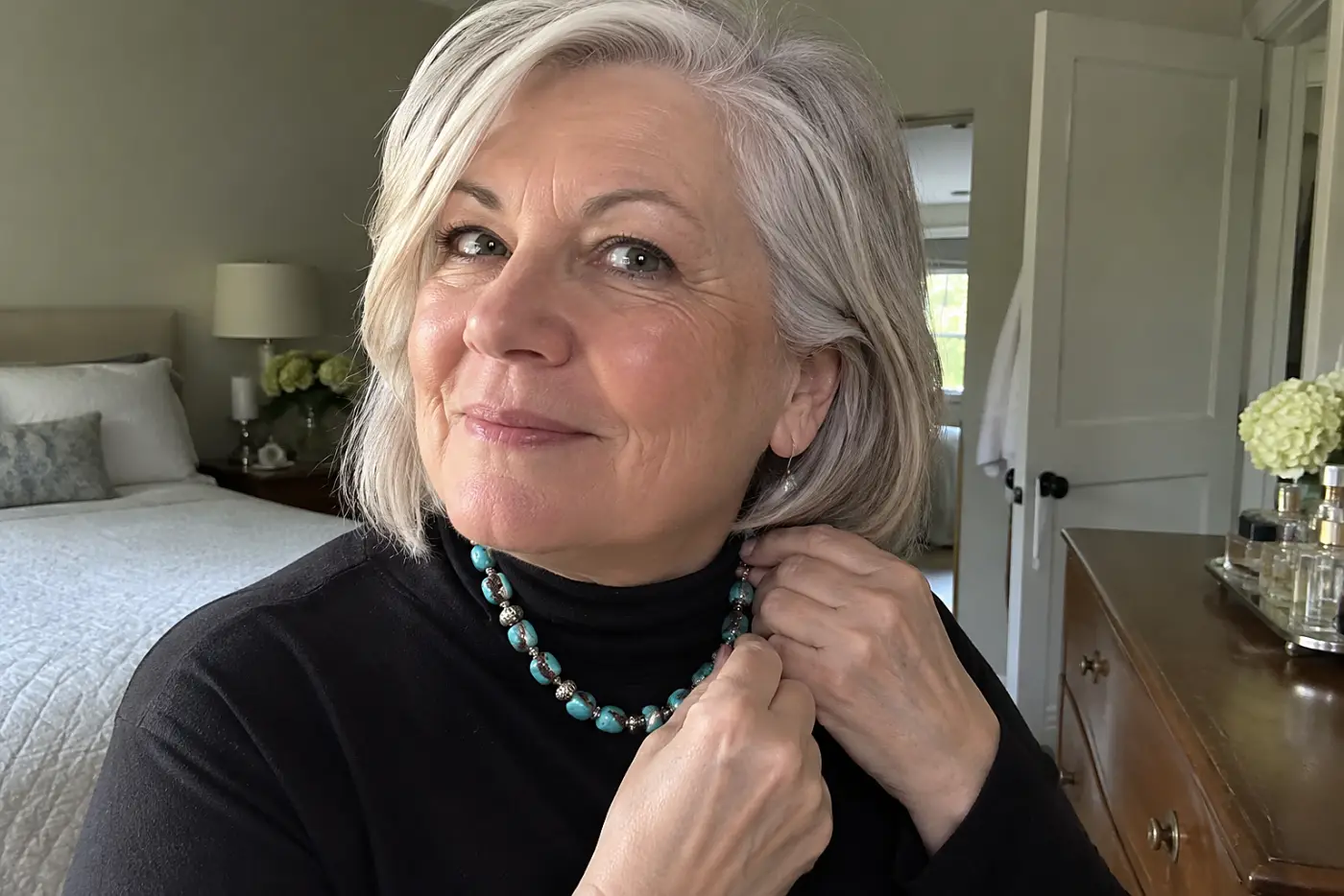 Older woman fastening a turquoise-and-silver necklace at her bedroom dresser.