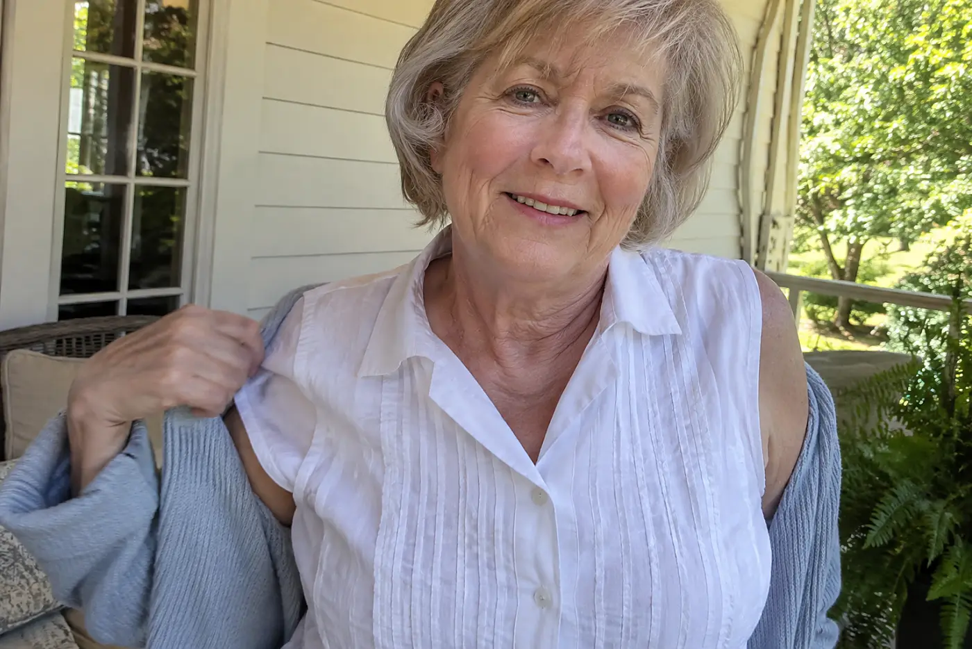 Older woman removing a cardigan over her cotton blouse on a warm porch.