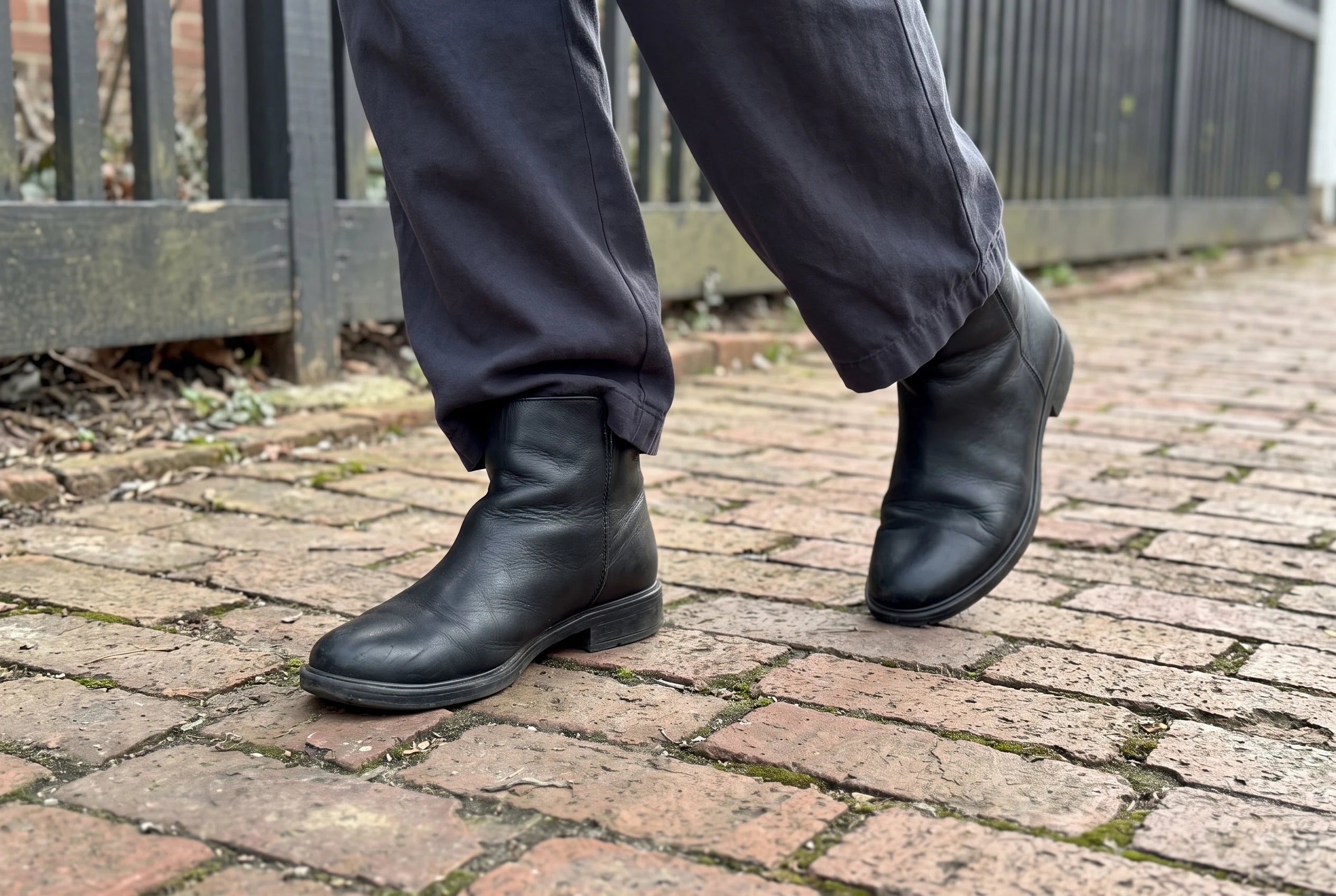 A woman wears black ankle boots and dark trousers while walking along a brick sidewalk.
