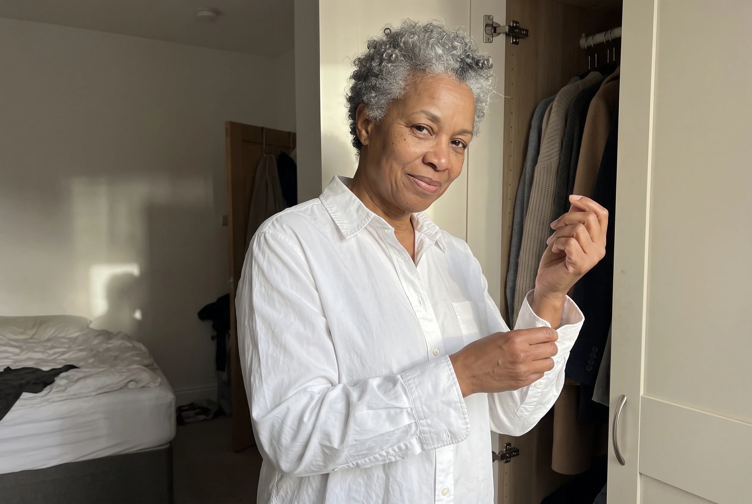 A woman in her sixties buttons a white cotton shirt beside her open wardrobe.