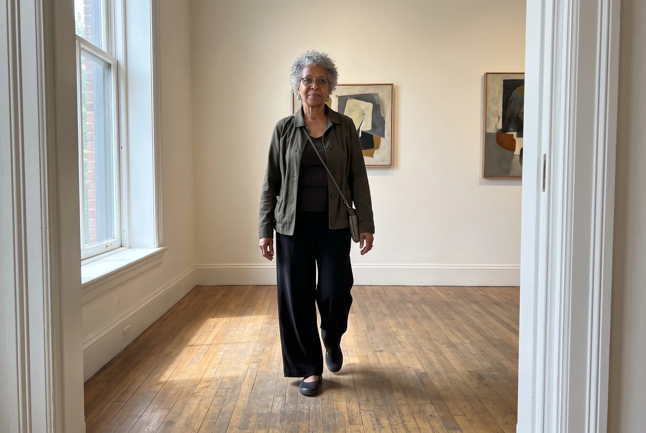 A woman in her sixties wearing black wide-leg trousers walks through an art gallery.