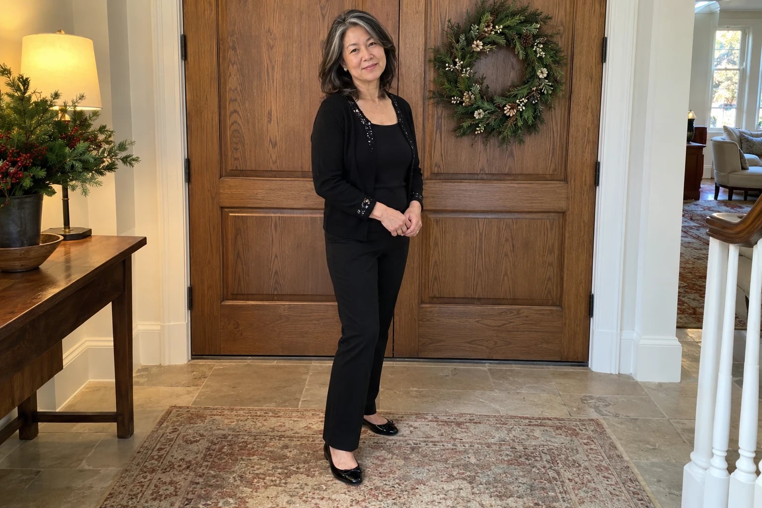A woman in her fifties stands by oak doors with a wreath in black pants and a sequin-trimmed cardigan.