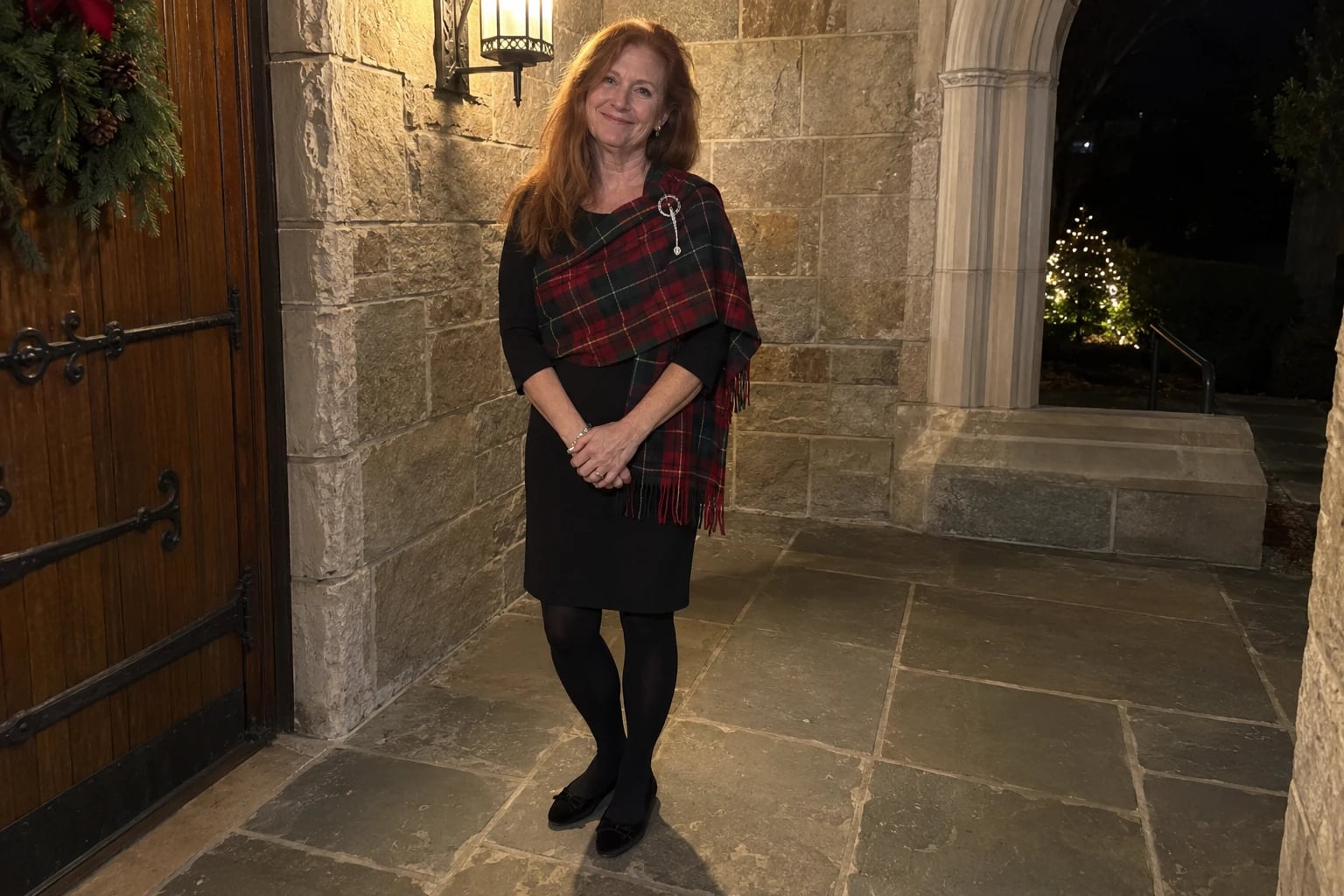 A woman in her fifties stands on a stone church porch wearing a black dress draped with a red tartan shawl.
