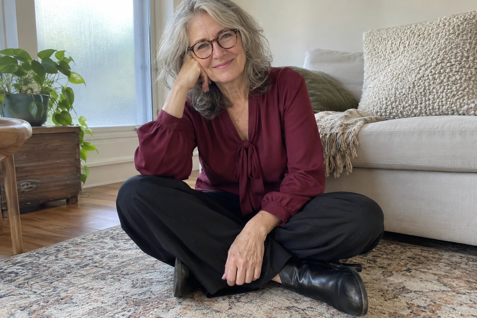 A woman in her sixties sits cross-legged on a rug in a garnet tie-neck blouse, black pants and ankle boots.