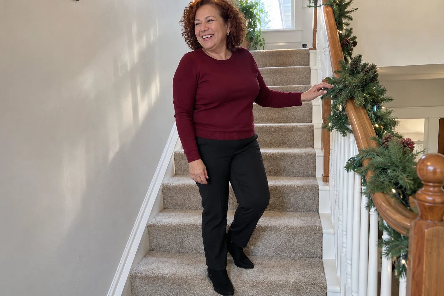 A woman in her sixties laughs descending a garland-wrapped staircase in a burgundy sweater, black pants and ankle boots.