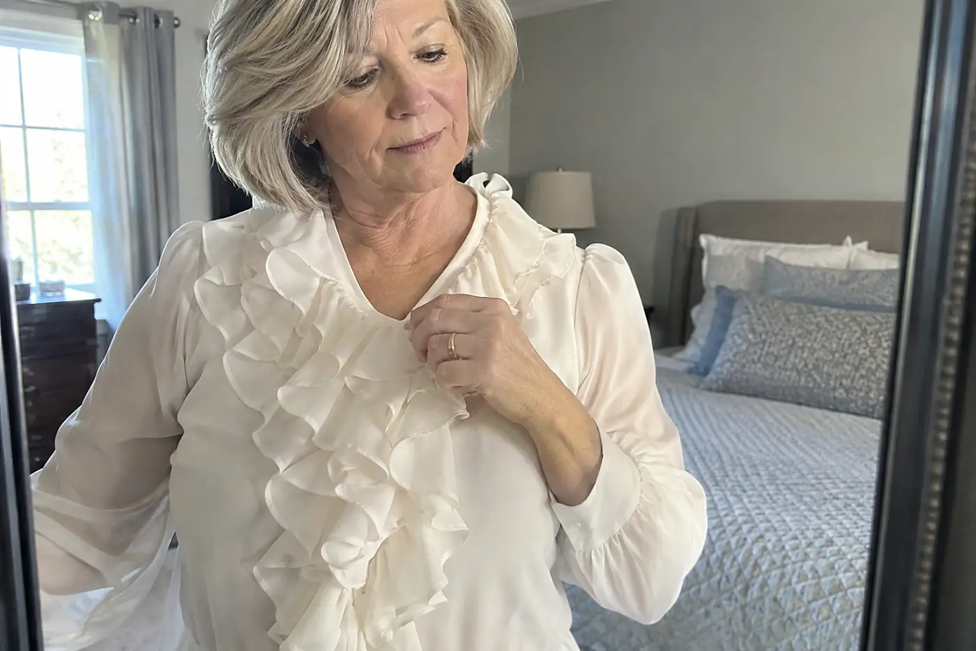 A woman in her sixties adjusts a white ruffled front blouse in a bedroom mirror.