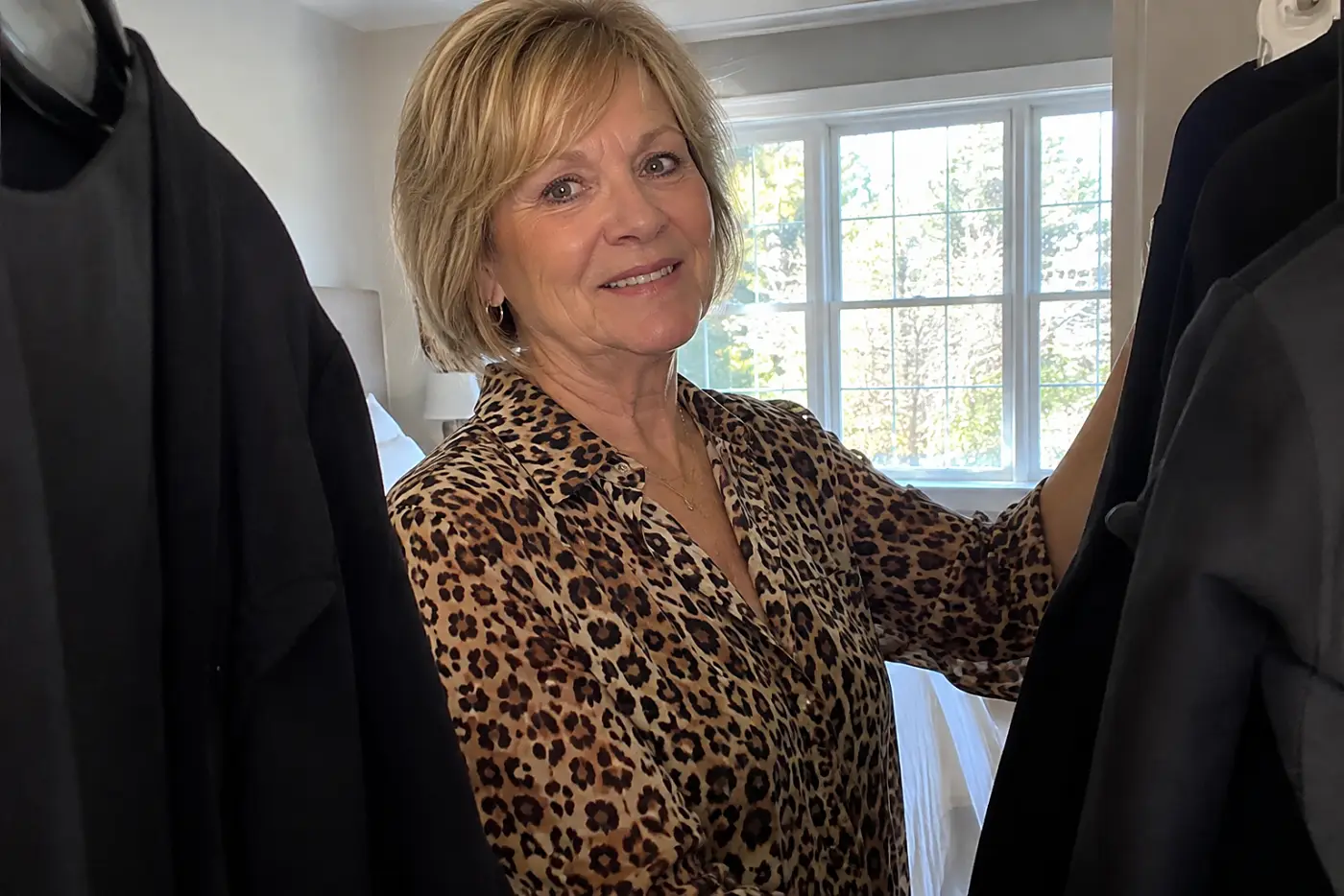 A leopard-print blouse is centered on a woman over 60 standing between two plain black jackets hanging in her bedroom.