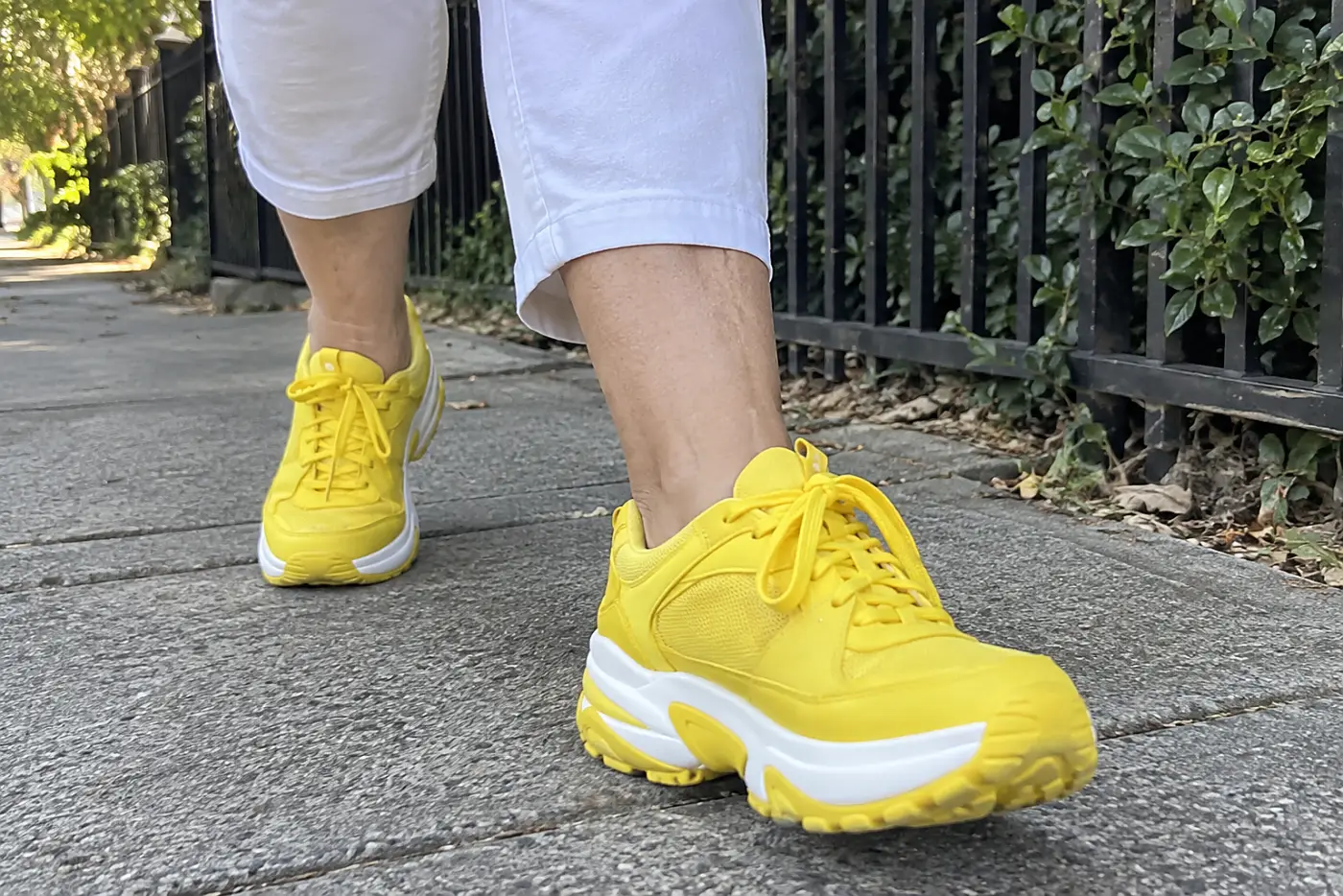 Bright yellow sneakers are centered on a woman over 60 walking along a gray neighborhood sidewalk.