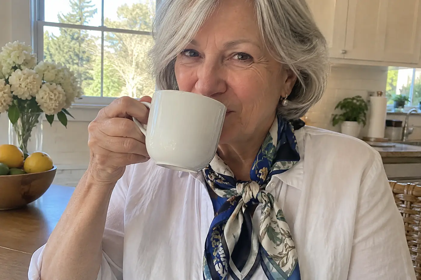 A silk scarf is centered at the collar of a woman over 60 drinking coffee at her kitchen table.