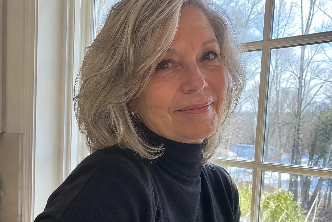 A black turtleneck is centered on a woman over 60 standing by her kitchen window in daylight.