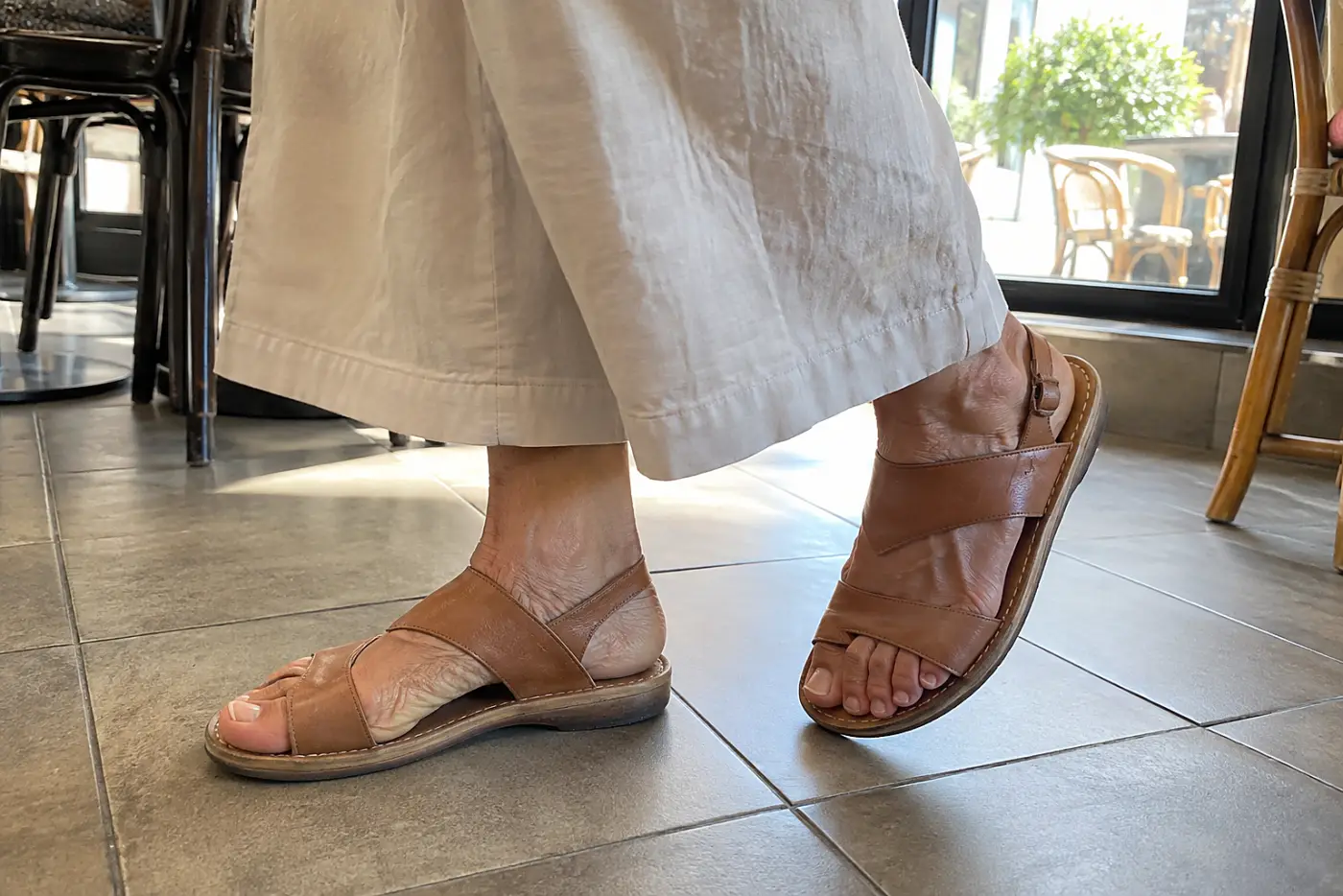 Flat leather sandals are centered on the feet of a woman over 60 crossing a tiled café floor.