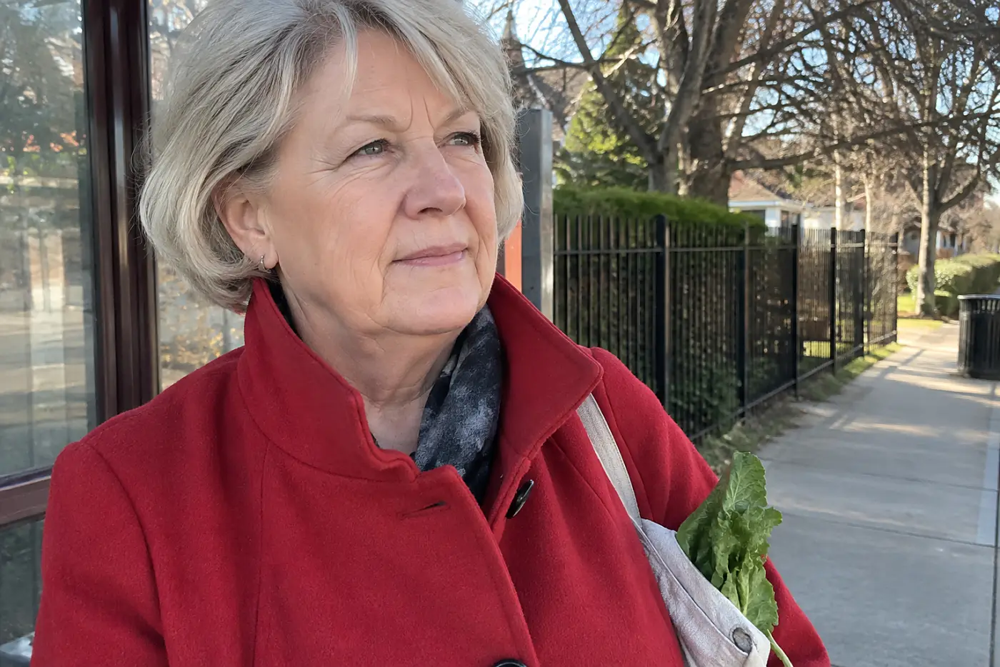 A woman over 60 in a red coat stands at a bus stop holding leafy greens.