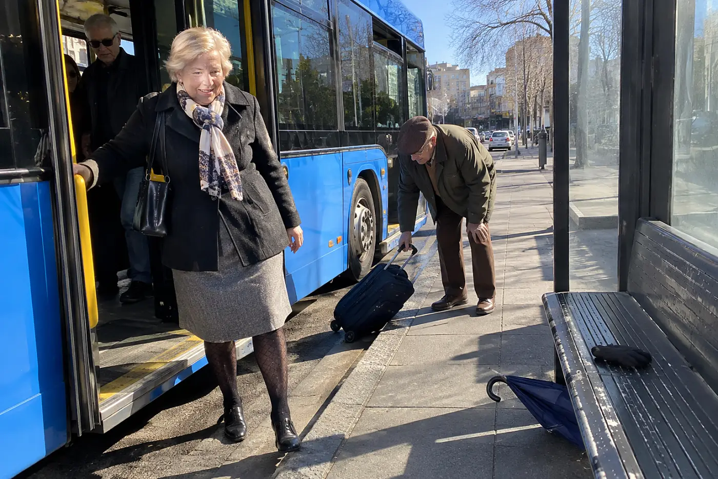 A woman in her sixties wearing patterned tights beneath a skirt steps off a bus.