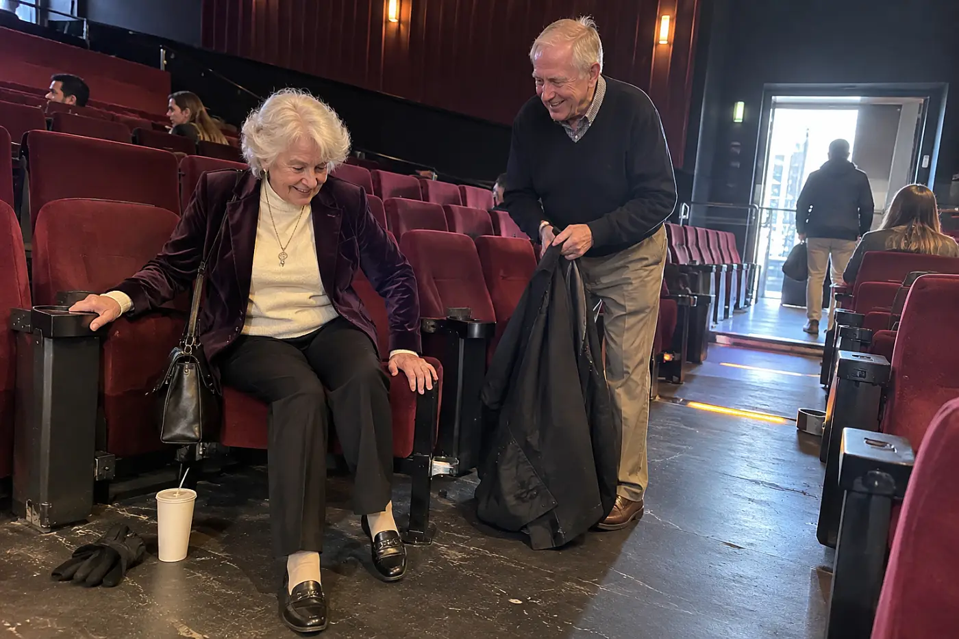 A woman in her seventies wearing a velvet blazer sits in a movie theater seat.