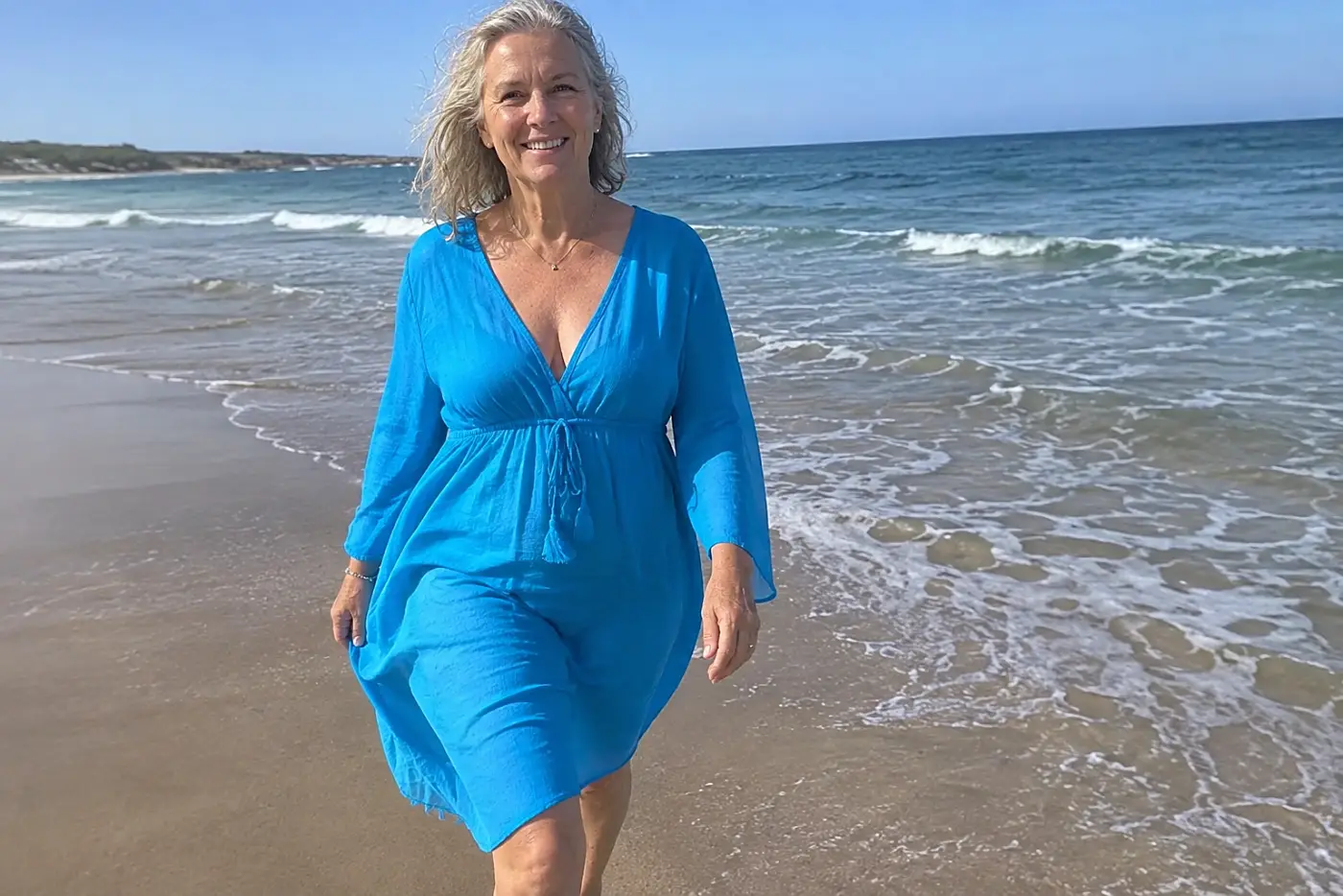 A bright blue swimsuit is centered on a woman over 60 walking toward the water at a beach.