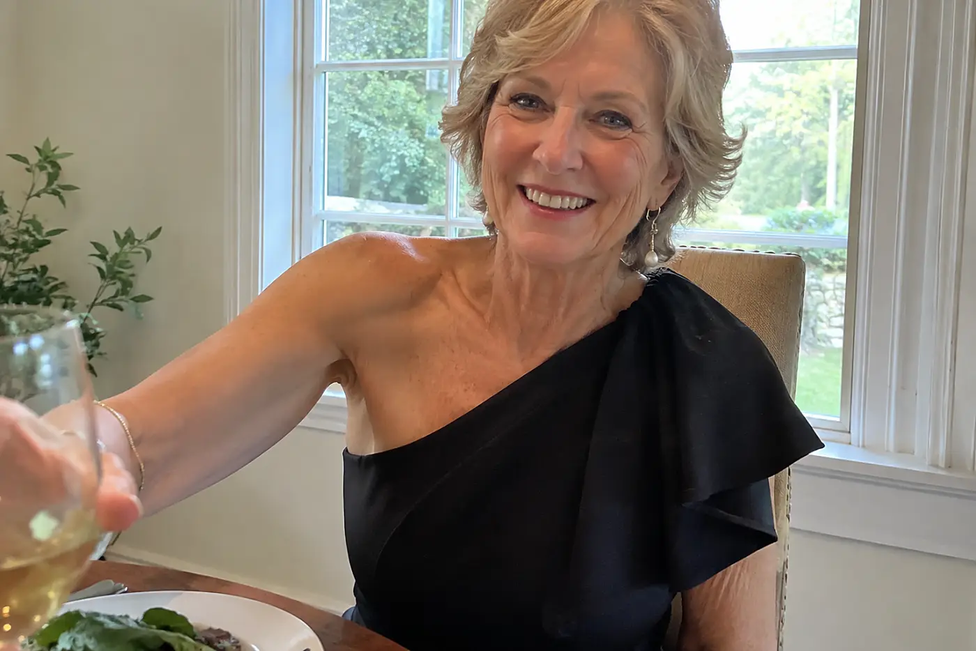 A one-shoulder top is centered on a woman over 60 reaching for a glass at a dinner table.