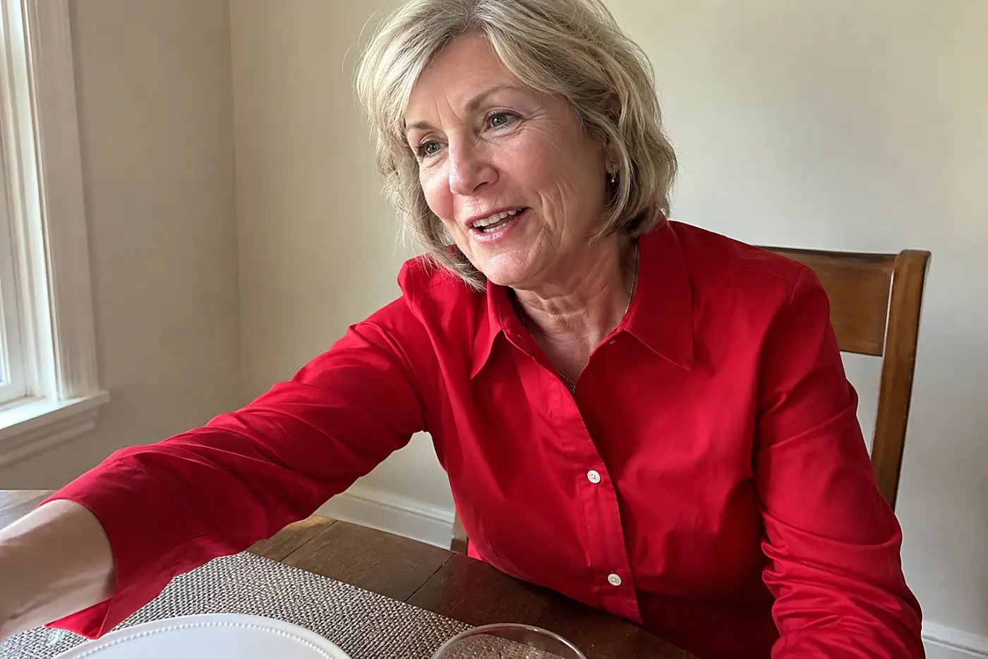 A woman in her sixties wears a bright red button-down shirt at a dining table.