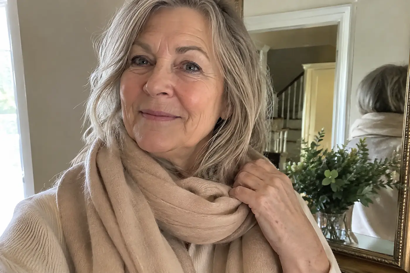 A woman in her sixties holds a beige scarf wrapped under her chin.