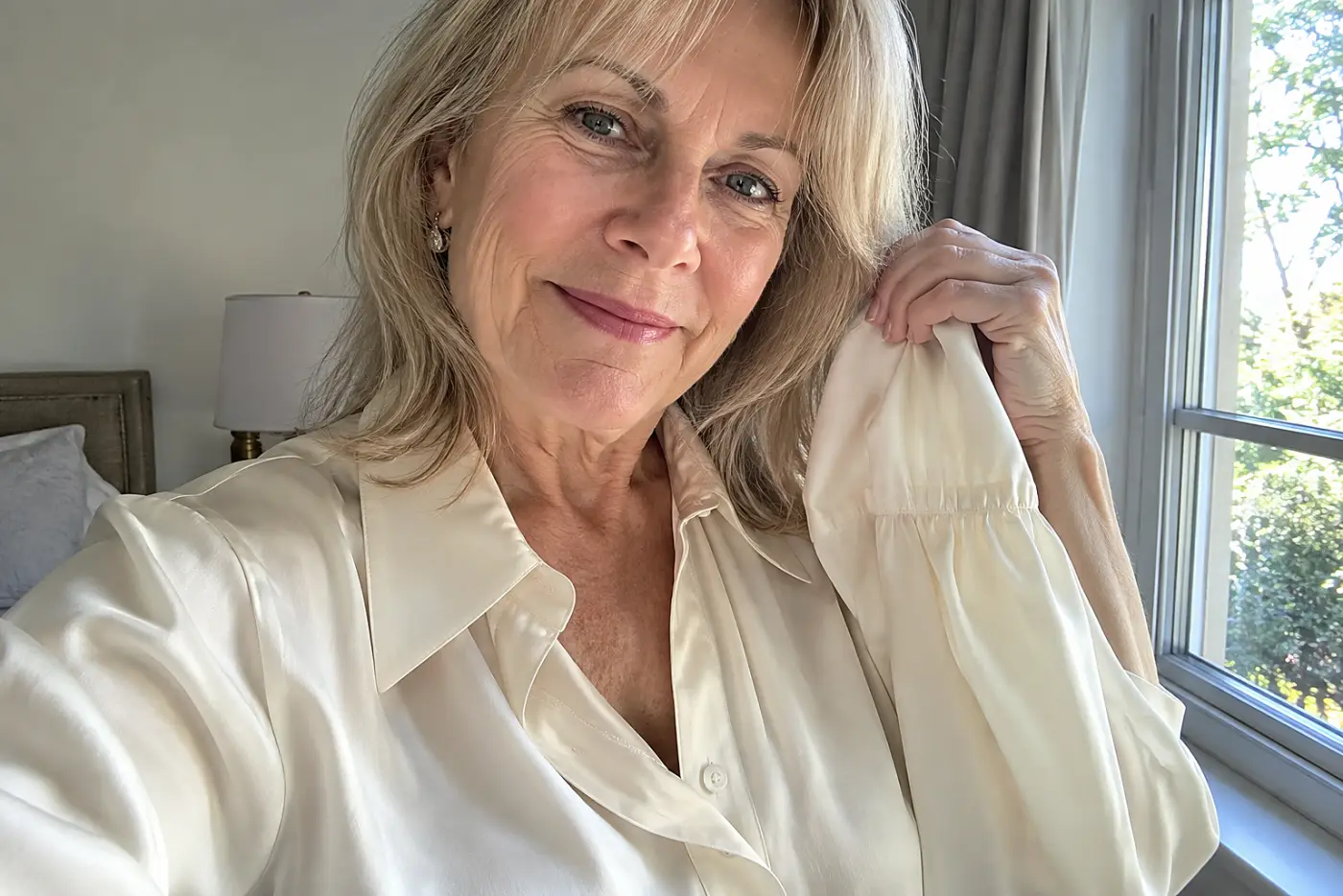 A blonde woman in her sixties wears a cream silk blouse near a bedroom window.