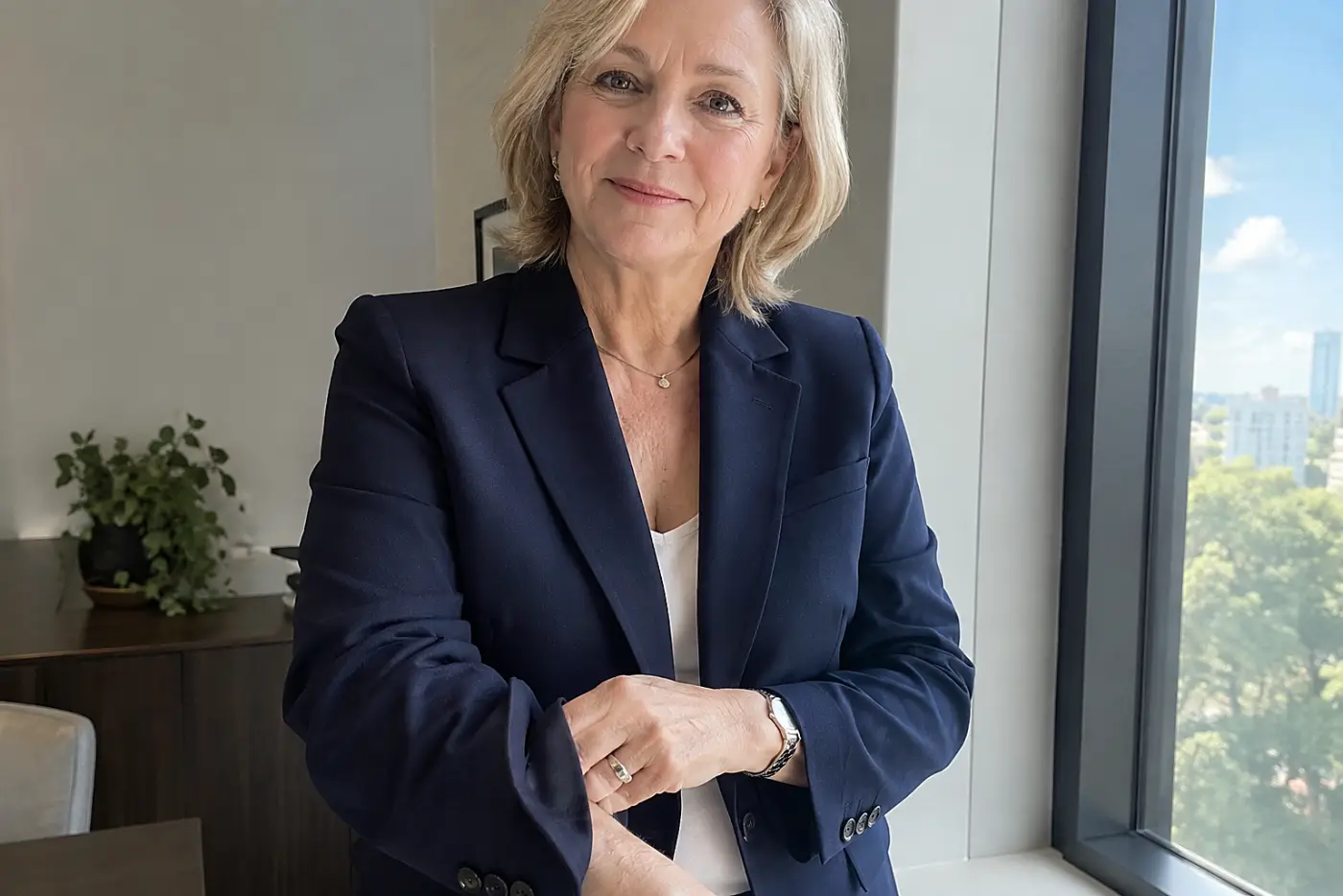 A woman in her sixties wears a navy blue blazer near a sunlit office window.