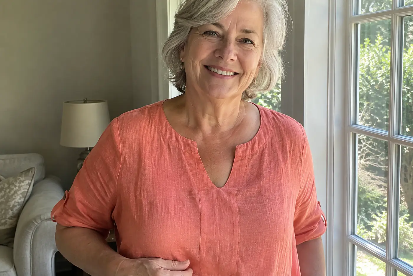 A smiling woman in her sixties wearing a coral linen top stands near a window.