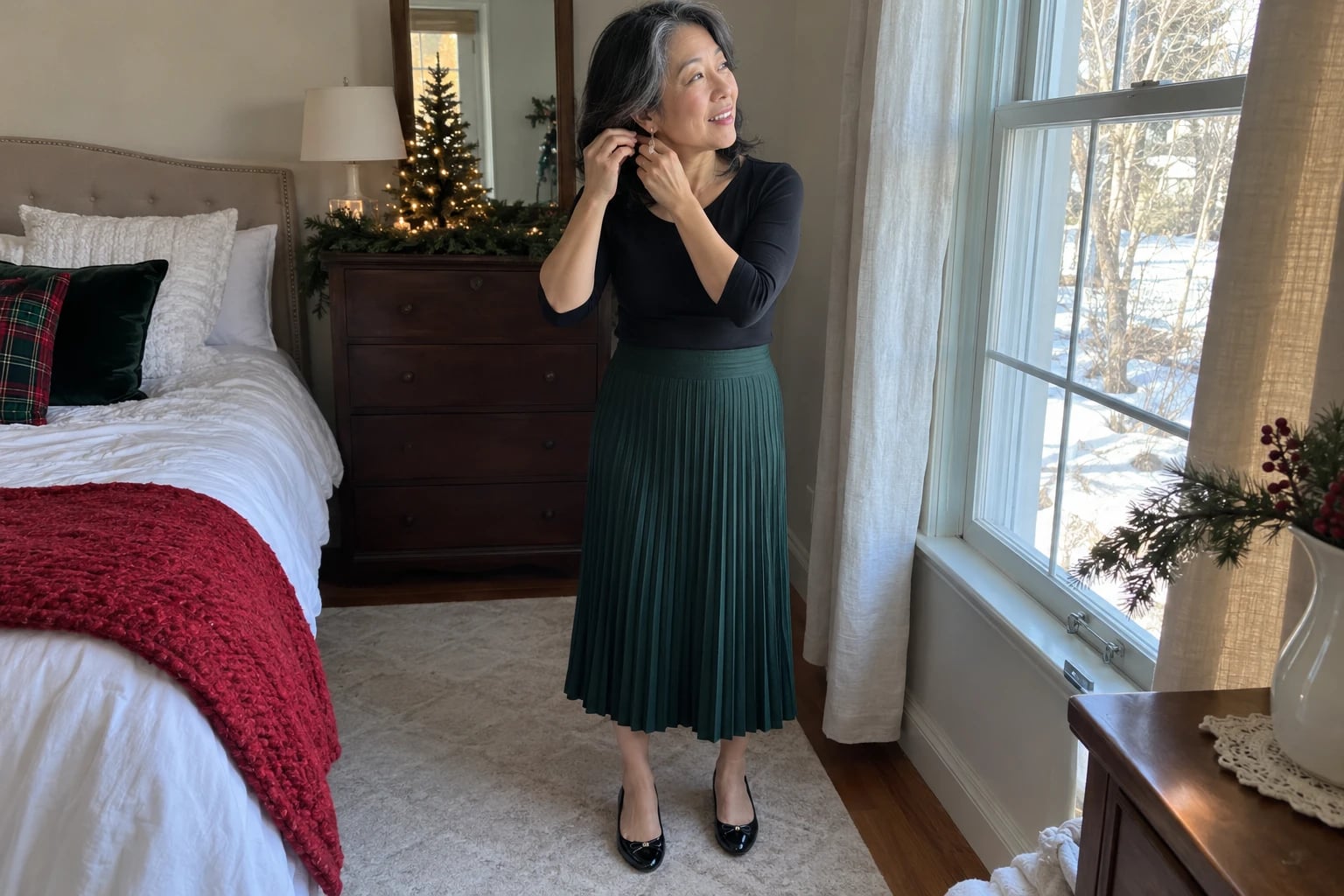 A woman in her fifties fastens an earring by a bedroom window, wearing a pine-green pleated midi skirt and black tee.