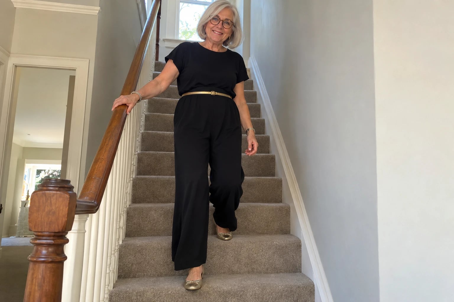 A woman in her sixties with silver bob and round glasses descends carpeted stairs in a black jumpsuit with gold belt and flats.
