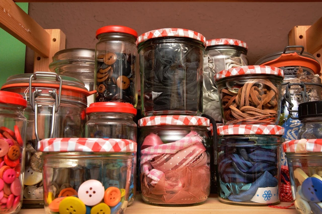Shelf of glass jars filled with colorful assorted buttons, alongside jars of ribbons and leather strips