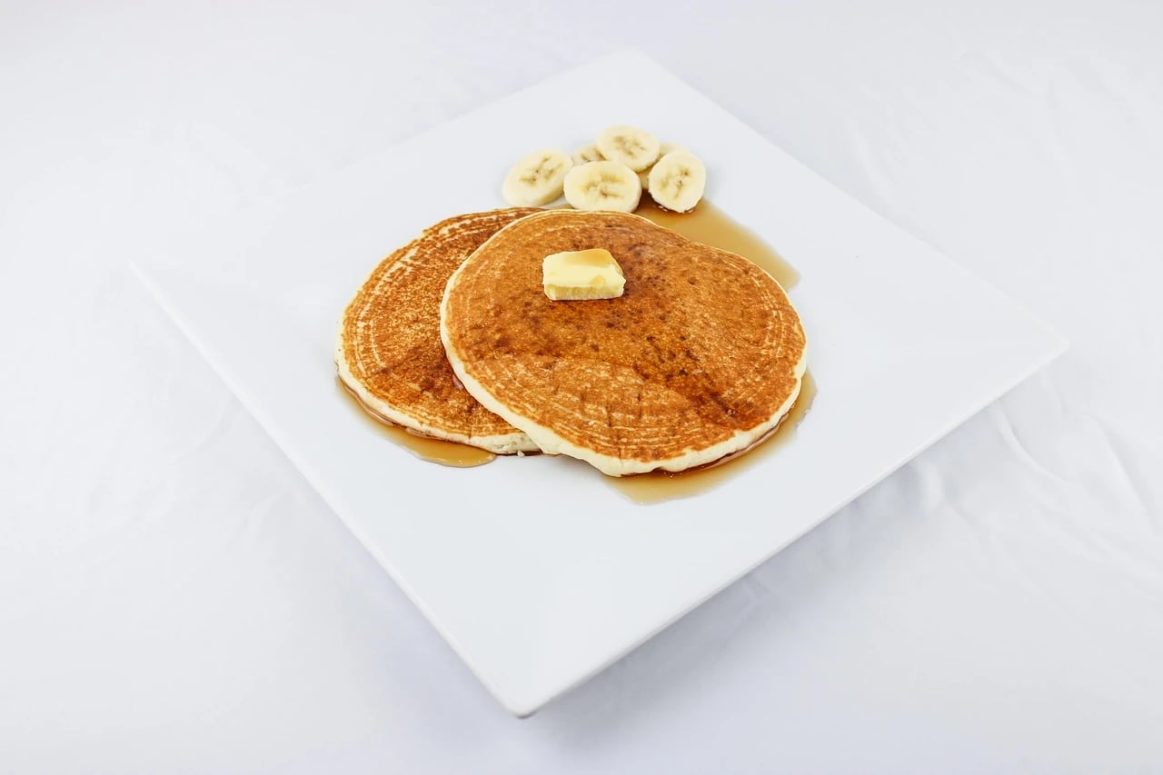 Two syrup-covered pancakes topped with butter and sliced bananas on a white square plate