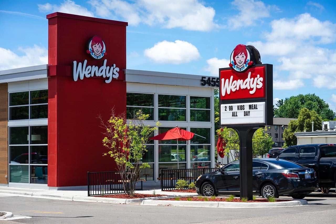 Wendy's restaurant exterior with red tower, logo signs, patio umbrella and a black car in the parking lot