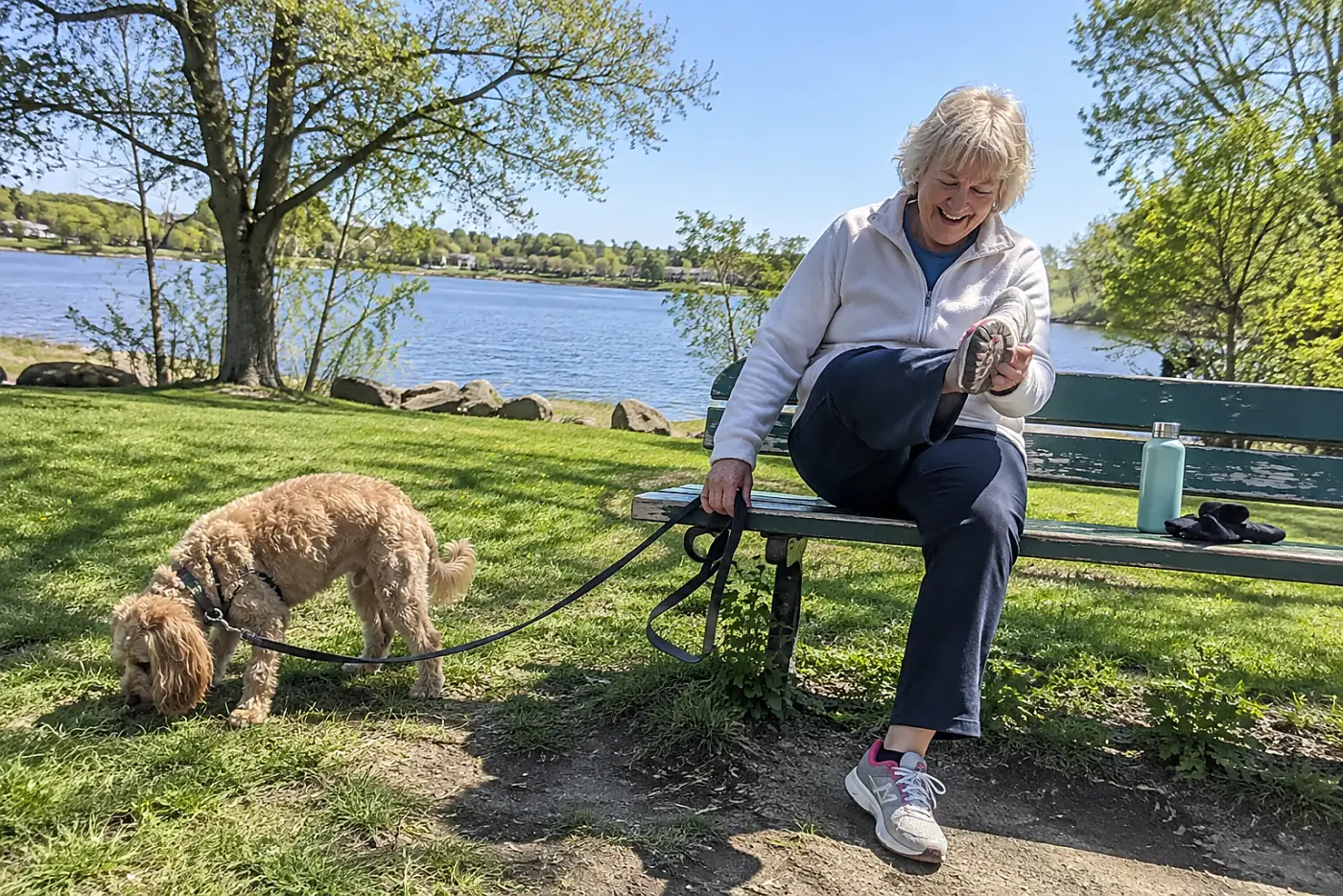 Older woman inspecting the sole of a shoe on a park bench while her dog sniffs nearby.