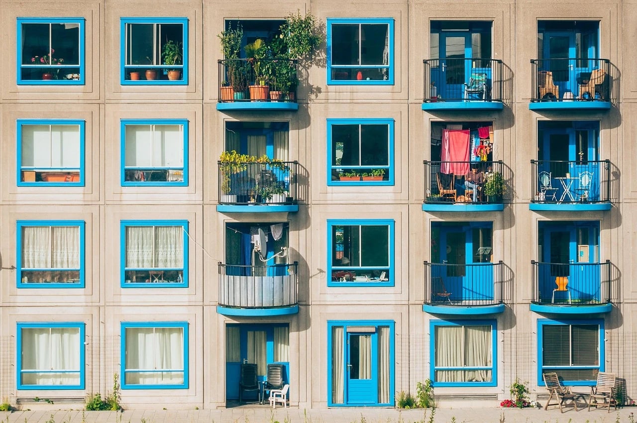 Apartment building facade with blue-framed windows and balconies holding chairs, tables, plants and hanging laundry