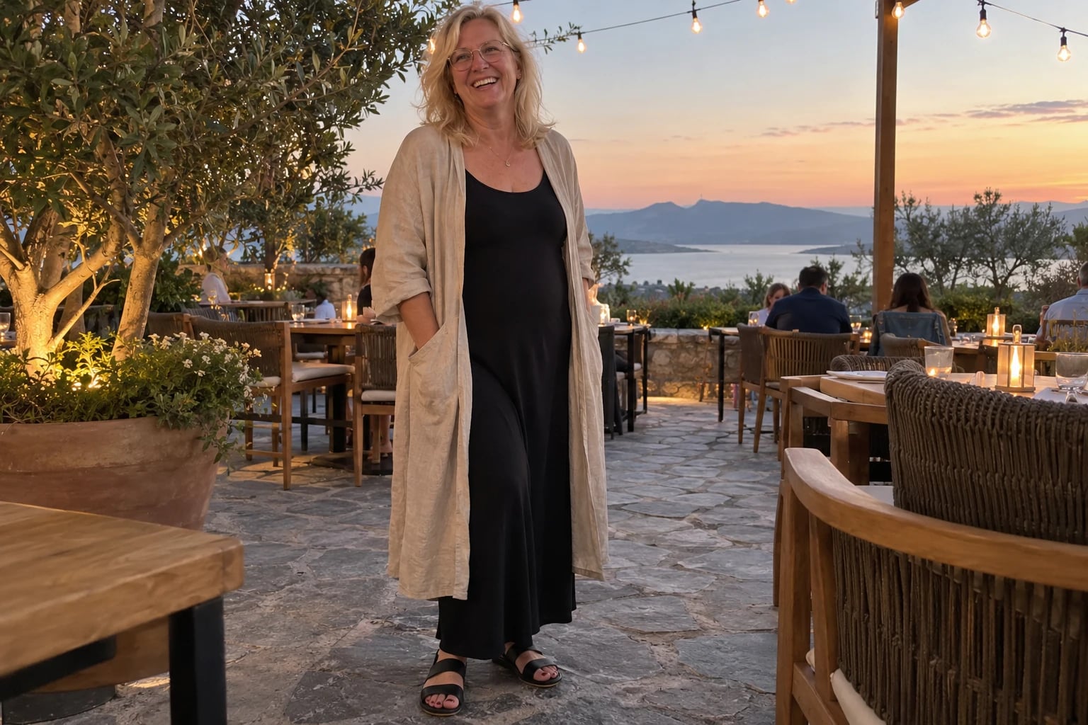 A woman over 50 smiles on a seaside patio in a black maxi dress with a beige linen duster and sandals.