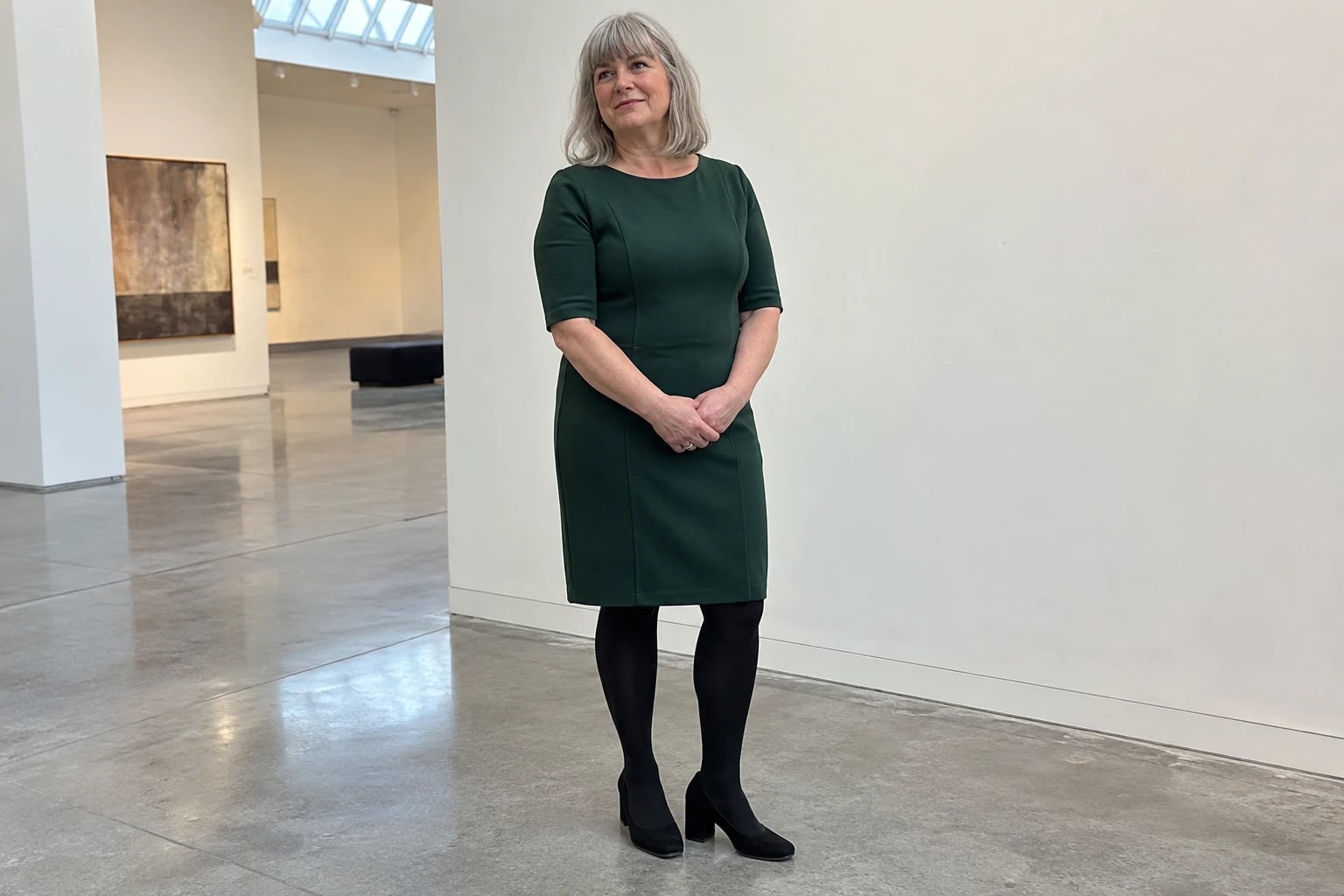 A gray-haired woman in her fifties stands in a museum gallery wearing a forest-green sheath dress with black tights and pumps.