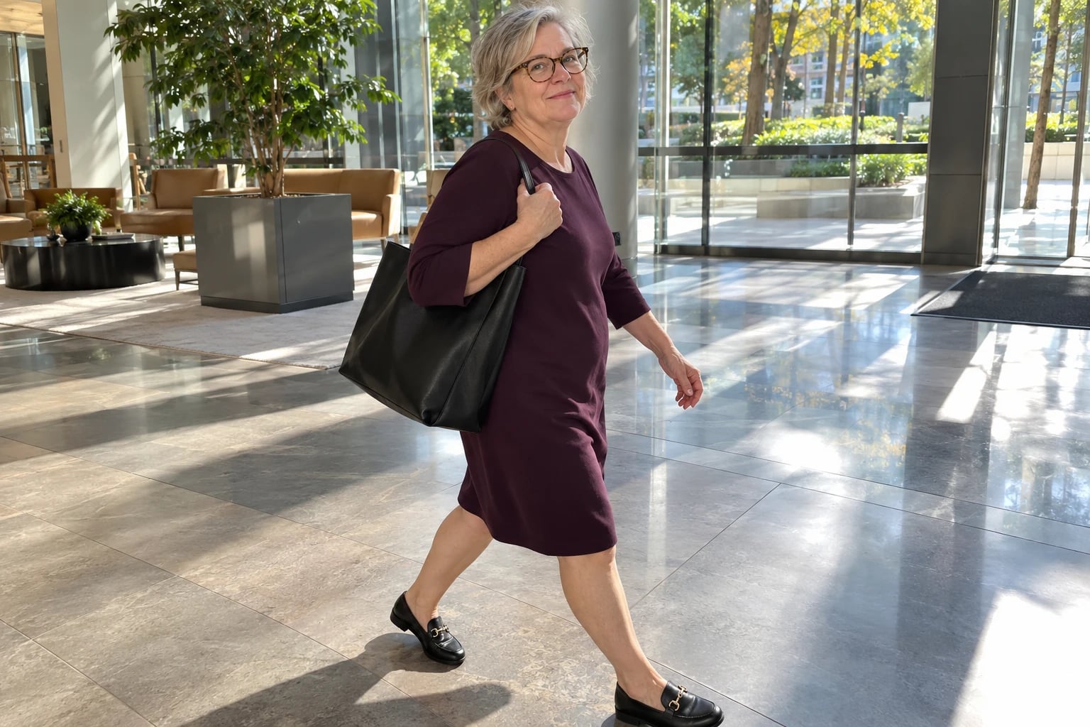 A gray-haired woman in round glasses walks through a sunlit lobby wearing a knee-length aubergine shift dress and black loafers.