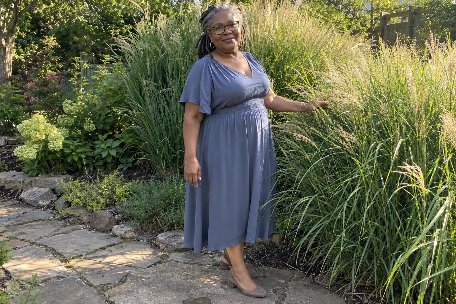A woman with gray locs and glasses wears a slate-blue empire waist dress beside tall ornamental grasses.