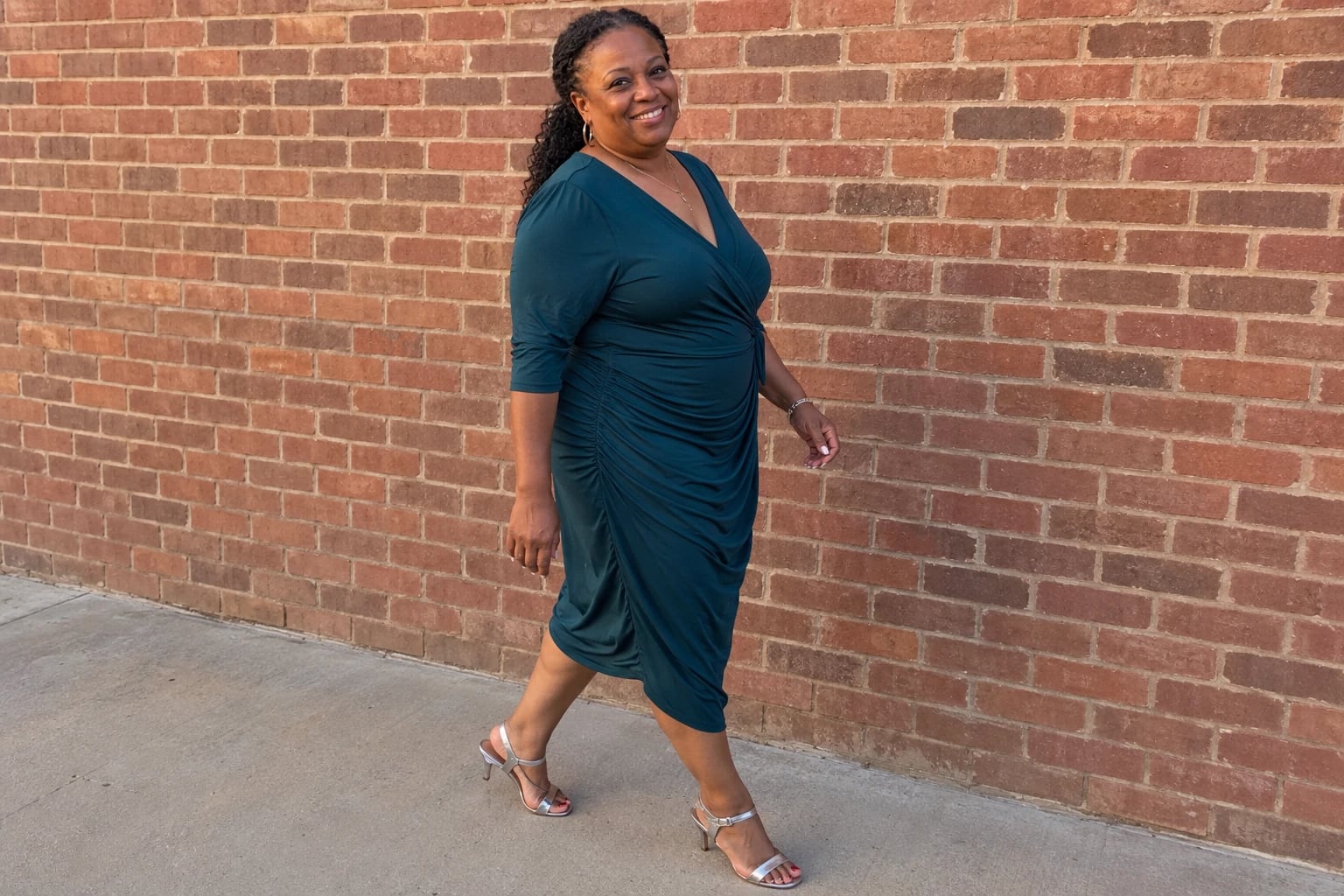 A woman in her fifties walks past a brick wall in a teal side-ruched wrap dress and silver sandals.