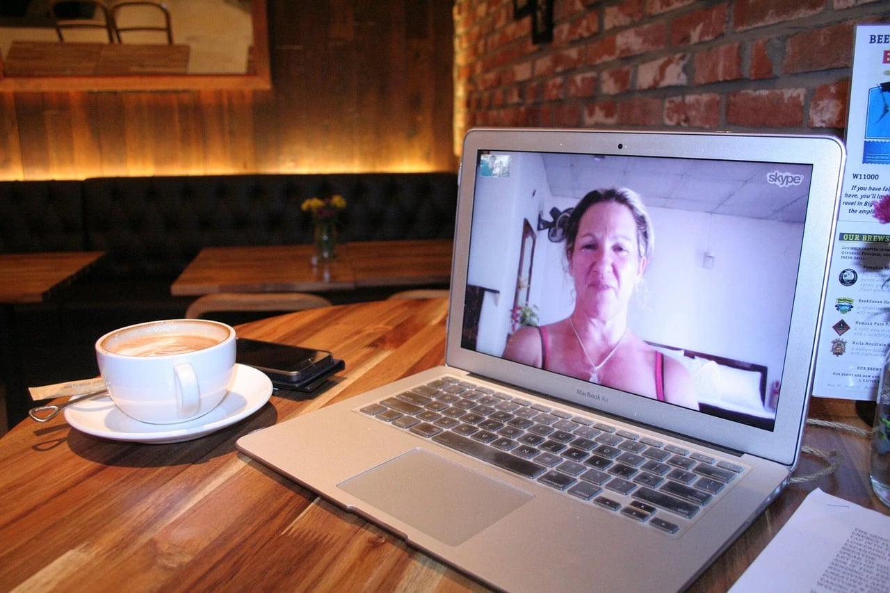 Middle-aged woman on a Skype video call displayed on a laptop at a cafe table beside a latte
